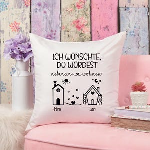 Könnte beinhalten: Weißes Kissen mit schwarzem Text, der "Ich wünschte, du würdest nebenan wohnen" lautet, mit zwei Häusern und den Namen "Nini" und "Lari" darunter.