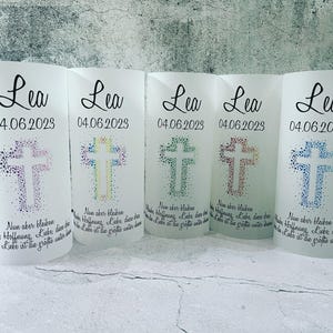 Könnte beinhalten: Fünf weiße, gefrostete Glas-Kerzenhalter mit dem Namen "Lea" und dem Datum "04.06.2023" auf jedem. Jeder Halter zeigt ein buntes Kreuz aus kleinen Punkten. Der Text "Nun aber bleiben" ist unter dem Kreuz auf jedem Halter gedruckt.