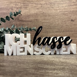 May include: A white block letter sign with the German phrase "Ich hasse Menschen" in black cursive script. The sign is on a wooden surface with greenery in the background.