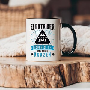 Könnte beinhalten: Weißer Keramikbecher mit schwarzem Henkel und Rand, mit dem Text "ELEKTRIKER HABEN ALLES ABER EINEN KURZEN" in Schwarz und Blau. Das Design beinhaltet ein Elektriker-Symbol. Der Becher steht auf einer Holzoberfläche.