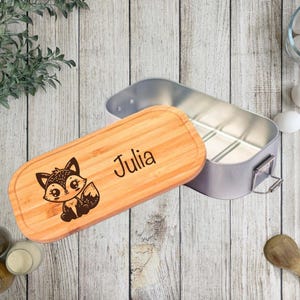 Könnte beinhalten: Eine personalisierte Brotdose aus Holz mit einem Cartoon-Fuchs-Design und dem Namen "Julia" auf dem Deckel eingraviert. Die Brotdose ist geöffnet und zeigt einen silbernen Innenraum. Der Deckel ist aus hellem Holz.