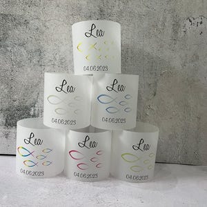 Könnte beinhalten: Sechs weiße Papier-Kerzenhalter mit dem Namen "Lea" und dem Datum "04.06.2023" auf jedem. Jeder Halter zeigt ein anderes Design mit bunten Fischen.
