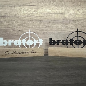 Puede incluir: Dos letreros de madera con la palabra "bratort" en blanco y negro, cada uno con un diseño de diana. El letrero blanco tiene grabado "Grillmeister Alex" en la base. Los letreros están sobre una superficie de madera.