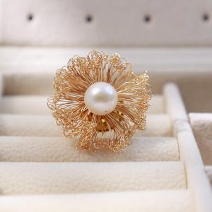 Puede incluir: Un anillo de alambre dorado con una perla blanca grande y redonda en el centro. El anillo tiene un diseño floral, con el alambre formando un delicado patrón calado. El anillo se exhibe en una caja de joyería blanca.