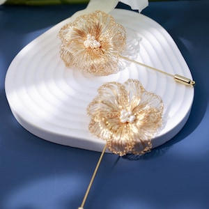 May include: Two gold-toned wire flower brooches with pearl centers rest on a white, petal-shaped dish. The brooches have long, thin pins. The background is a deep blue surface with white flowers.