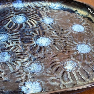 Puede incluir: Una bandeja de cerámica con base marrón oscuro y un estampado floral en tonos azules. La bandeja tiene una forma irregular y orgánica y un acabado brillante. Las flores están en relieve.