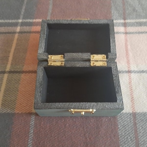 Op de afbeelding: Kleine, open, grijze houten kist met een zwart interieur. De kist heeft goudkleurige scharnieren en een sluiting. De kist staat op een geruit oppervlak.
