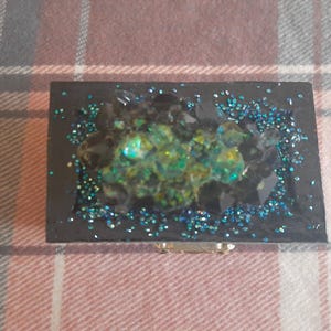 Op de afbeelding: Een rechthoekige zwarte sieradendoos met een geode-achtig ontwerp. Het midden heeft een cluster van groene en gele kristallen, omringd door zwarte stenen en blauwe glitter. Een kleine, goudkleurige sluiting is zichtbaar.