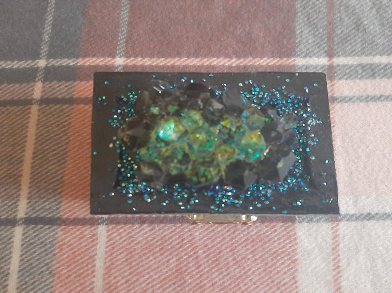 Op de afbeelding: Een rechthoekige zwarte sieradendoos met een geode-achtig ontwerp. Het midden heeft een cluster van groene en gele kristallen, omringd door zwarte stenen en blauwe glitter. Een kleine, goudkleurige sluiting is zichtbaar.