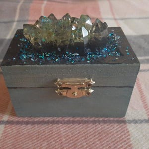 A cool looking dark teal trinket box with hints of silver and blue glitter dry brushed on the box. Embellished with an iridescent resin crystal and smaller iridescent blue crystals surrounding it. The inside is painted black with pearlescent paint on the rim to hide your mysterious treasures. It will add a pop of fantasy and mystery to any space.