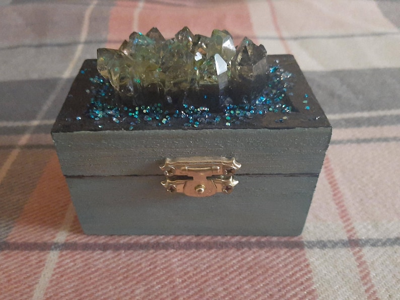 A cool looking dark teal trinket box with hints of silver and blue glitter dry brushed on the box. Embellished with an iridescent resin crystal and smaller iridescent blue crystals surrounding it. The inside is painted black with pearlescent paint on the rim to hide your mysterious treasures. It will add a pop of fantasy and mystery to any space.