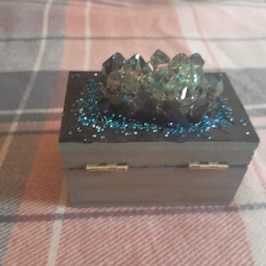 Op de afbeelding: Een kleine, houten doos met scharnieren en een donkergrijze afwerking. De bovenkant is versierd met blauwe glitter en een cluster van heldere, facetgeslepen kristallen. De doos heeft goudkleurige scharnieren.