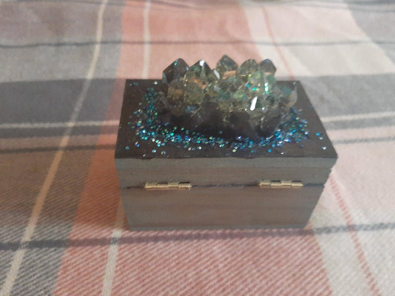 Op de afbeelding: Een kleine, houten doos met scharnieren en een donkergrijze afwerking. De bovenkant is versierd met blauwe glitter en een cluster van heldere, facetgeslepen kristallen. De doos heeft goudkleurige scharnieren.