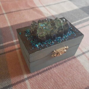 Op de afbeelding: Een kleine, rechthoekige, grijze houten doos met een goudkleurige sluiting. De bovenkant is versierd met blauwe glitter en een cluster van iriserende, kristalachtige formaties. De doos is waarschijnlijk voor sieraden of kleine aandenkens.