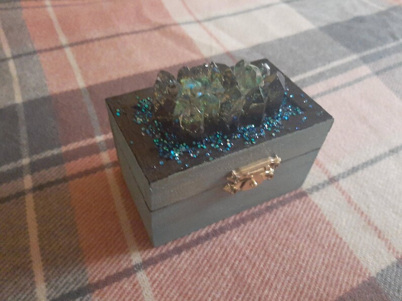 Op de afbeelding: Een kleine, rechthoekige, grijze houten doos met een goudkleurige sluiting. De bovenkant is versierd met blauwe glitter en een cluster van iriserende, kristalachtige formaties. De doos is waarschijnlijk voor sieraden of kleine aandenkens.