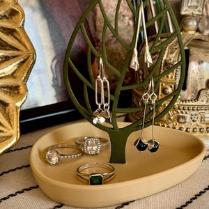 Op de afbeelding: Een groene, blaadjesvormige juwelendoos met een beige schaal. De doos heeft drie paar oorbellen en een ringenschaal met drie ringen. De ringen zijn zilverkleurig met heldere en groene stenen.