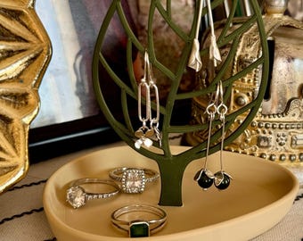 May include: A green leaf-shaped jewelry holder with a beige dish. The holder has three pairs of earrings and a ring dish with three rings. The rings are silver with clear and green stones.