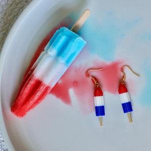 May include: A red, white, and blue popsicle with a wooden stick rests on a white plate, next to a pair of popsicle-shaped earrings. The earrings have gold-colored hooks and are colored to match the popsicle.