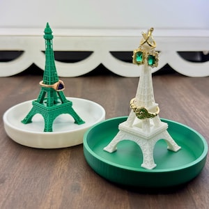 May include: Two Eiffel Tower-shaped ring holders. One is green with a white base, the other is white with a green base. Each holds rings and earrings. The holders sit on a wooden surface.