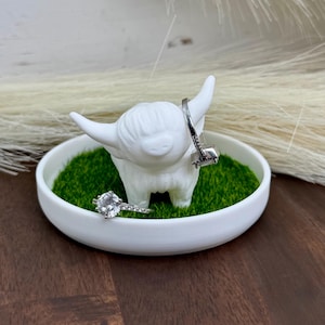 Könnte beinhalten: Weißer Keramik-Ringhalter in Form eines Hochlandrindes mit grünem Grasuntergrund. Zwei silberne Ringe werden auf dem Halter präsentiert.