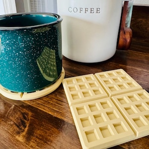May include: A teal speckled enamel mug with a waffle design coaster, alongside a cream-colored coffee canister. Four square waffle-shaped coasters are arranged on a wooden surface. The canister has the word "COFFEE" printed on it.