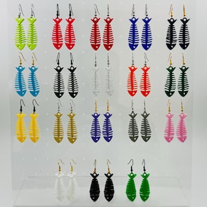 Puede incluir: Exhibición de pendientes con forma de espina de pescado en varios colores, incluyendo verde, rojo, azul, negro, blanco, amarillo, dorado, morado, gris y rosa. Cada par cuelga de un gancho plateado o dorado.