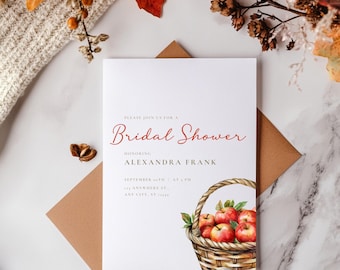 Puede incluir: Una tarjeta de invitación blanca con el texto "Bridal Shower" en escritura roja. La tarjeta presenta una ilustración de acuarela de una cesta llena de manzanas rojas. La invitación está en un sobre marrón.