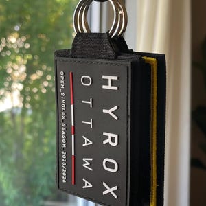 May include: A black rectangular keychain featuring the white text HYROX OTTAWA and the vertical text OPEN_SINGLES_SEASON_2025/2026. The keychain has a red accent line, a yellow stitched edge, and is attached to a silver metal split ring.