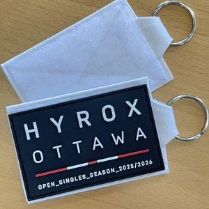 以下が含まれることがあります： 銀色の金属リングが付いた白い布製の長方形キーホルダー2つ。1つには、白い文字でHYROX OTTAWA、赤と白の線、OPEN_SINGLES_SEASON_2025/2026という文字が書かれた黒いゴム製のパッチが付いています。もう1つは無地の白い布製です。