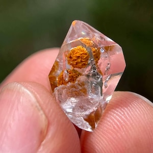 May include: A clear quartz crystal with orange inclusions, held between fingers. The crystal is faceted with a pointed top and contains several spherical, orange-colored formations. The background is a blurred green.