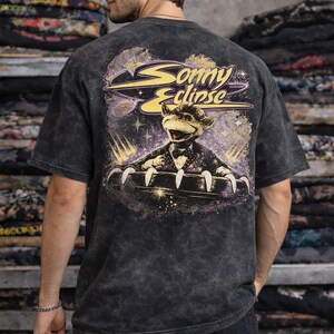 May include: Dark gray t-shirt with a graphic on the back featuring a cartoon character in a tuxedo with the text "Sonny Eclipse". The design includes stars and a planet. The shirt is worn by a person.