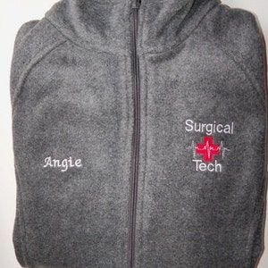 Puede incluir: Chaqueta de forro polar gris con cremallera y cuello alto. La chaqueta tiene el texto bordado "Angie" en el pecho izquierdo y "Surgical Tech" con un diseño de cruz roja en el pecho derecho. La cremallera es gris oscura.