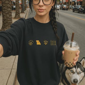 May include: Black sweatshirt with embroidered gold and yellow designs. The designs include a paw print with an "M", a yellow state outline with a red heart, a tiger head, and four vertical lines. The person is holding a bubble tea and walking a husky.