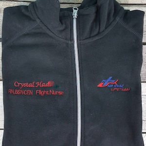 Puede incluir: Chaqueta negra de forro polar con cremallera y cremallera gris y texto bordado en el lado izquierdo: "Crystal Had RN, BSN, CEN Flight Nurse" y en el lado derecho: "AIR EVAC LIFETEAM".