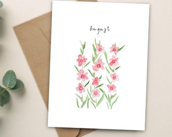 Könnte beinhalten: Eine weiße Grußkarte mit einer Aquarellillustration von rosa Blumen mit grünen Stielen und Blättern. Das Wort August ist in schwarzer Schrift über dem Blumenarrangement gedruckt. Ein brauner Umschlag liegt hinter der Karte.