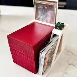 May include: A white card holder containing a red deck of cards, a card with text, and a green die. The card holder has slots for cards and a die. The card in the holder has the text "Stefan, Maurer Progenitor".