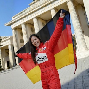 May include: A person in a red racing suit holds a German flag in front of the Brandenburg Gate. The flag is black, red, and yellow. The racing suit has the Marlboro logo.