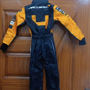 May include: A black and orange McLaren Formula 1 team racing suit. The suit has the McLaren logo on the back and sponsor logos on the sleeves. The suit is hanging on a metal hanger.