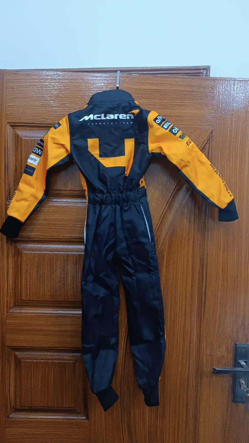 May include: A black and orange McLaren Formula 1 team racing suit. The suit has the McLaren logo on the back and sponsor logos on the sleeves. The suit is hanging on a metal hanger.