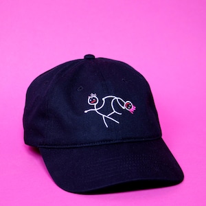 Peut inclure: Une casquette de baseball noire avec un motif brodé blanc de deux personnages en bâton. Les personnages se font face, l'un tenant la main de l'autre.
