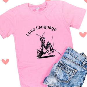 May include: A pink t-shirt with the words "Love Language" and a black praying mantis graphic. The shirt is paired with denim shorts. Pink hearts are scattered around the image.