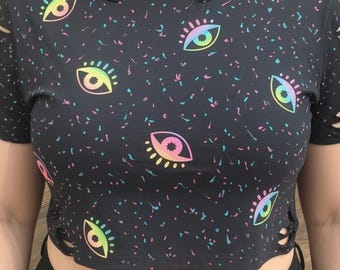 May include: A black short-sleeved crop top featuring a repeating pattern of iridescent, rainbow-colored eyes and small colorful speckles. The garment includes distressed, ripped detailing along the neckline and sleeves, with side lace-up accents.
