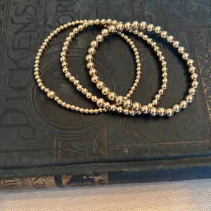 May include: Three gold beaded bracelets of varying sizes are arranged on a dark blue, textured surface. The bracelets are made of small, round, shiny gold beads, creating a delicate and elegant look. The bracelets are a simple yet stylish accessory.