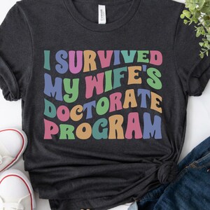 Ich habe das Doktorandenprogramm meiner Frau überlebt, Frau Doktoranden-Shirt, lustiges PHD-Abschlussgeschenk, Ehemann-Stützen-T-Stück, PHD Party 2026 T-Shirt