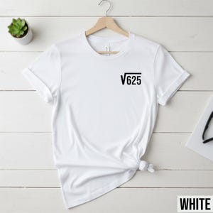 May include: White t-shirt with the black text "√625" printed on the front. The shirt is displayed on a wooden hanger against a white wooden background. A small potted plant is visible in the upper left corner.