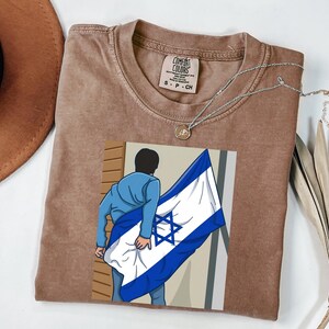 May include: A brown Comfort Colors t-shirt with a graphic of a person holding the Israeli flag. A silver necklace with a small pendant is on the shirt. A brown hat and a dried palm leaf are in the background.