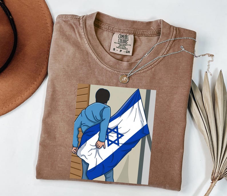 May include: A brown Comfort Colors t-shirt with a graphic of a person holding the Israeli flag. A silver necklace with a small pendant is on the shirt. A brown hat and a dried palm leaf are in the background.