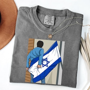 May include: A gray Comfort Colors t-shirt featuring a graphic of a person holding the Israeli flag. The shirt has a crew neck and short sleeves. A gold necklace is draped across the shirt.