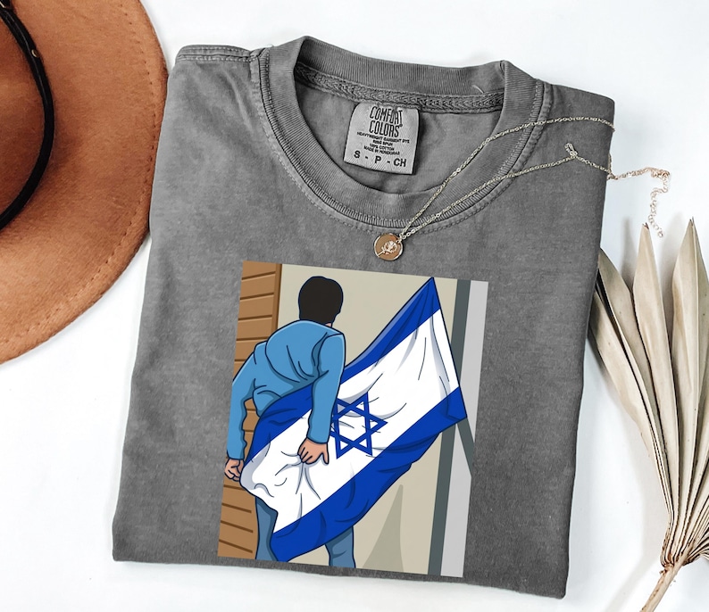 May include: A gray Comfort Colors t-shirt featuring a graphic of a person holding the Israeli flag. The shirt has a crew neck and short sleeves. A gold necklace is draped across the shirt.
