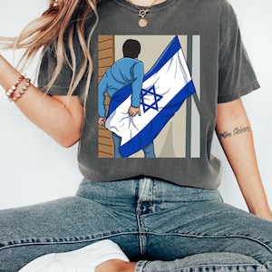 May include: Dark grey t-shirt featuring a graphic of a person holding the Israeli flag. The flag is white with a blue Star of David and blue stripes. The person is wearing a blue jacket and jeans. The shirt is a casual style.